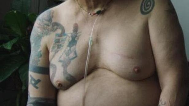 Eine tätowierte, ältere Person mit weißem Haar und einem Nasenkatheter sitzt.