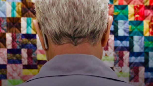 Ein Mann betrachtet von hinten ein farbenfrohes Patchwork-Muster, das an eine Steppdecke erinnert; Filmposter „The Quilters“.