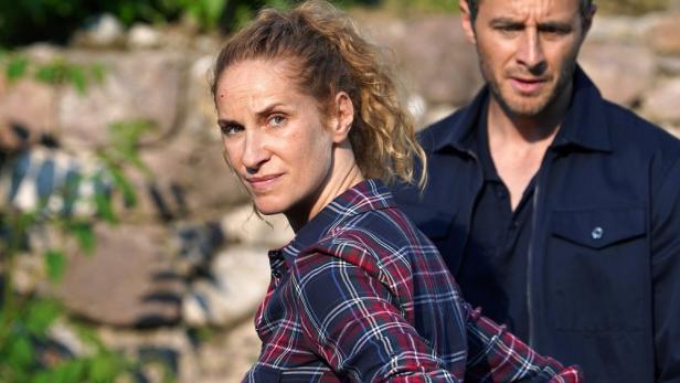 Zwei Schauspieler, eine Frau im karierten Hemd und ein Mann in einer blauen Jacke, stehen vor einer Steinmauer.