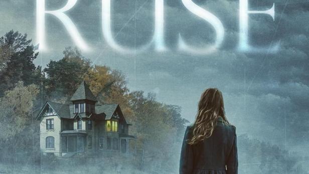 Eine Frau mit Koffer steht am Ufer und blickt auf ein abgelegenes Haus am Wasser.