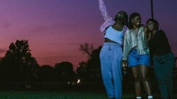 Drei junge Frauen stehen bei Dämmerung auf einer Wiese, eine hält ihr Handy in die Luft.