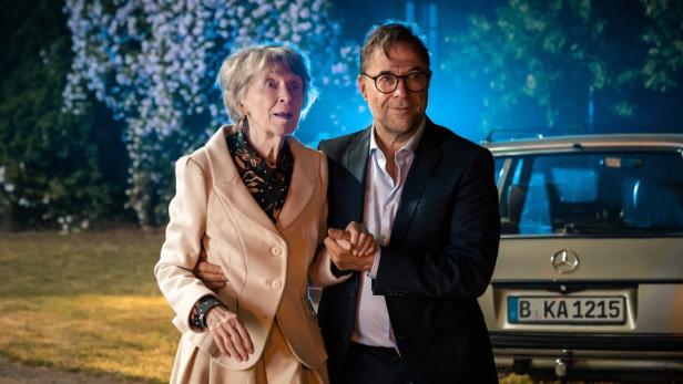 Eine ältere Frau und ein Mann halten Händchen vor einem Mercedes.