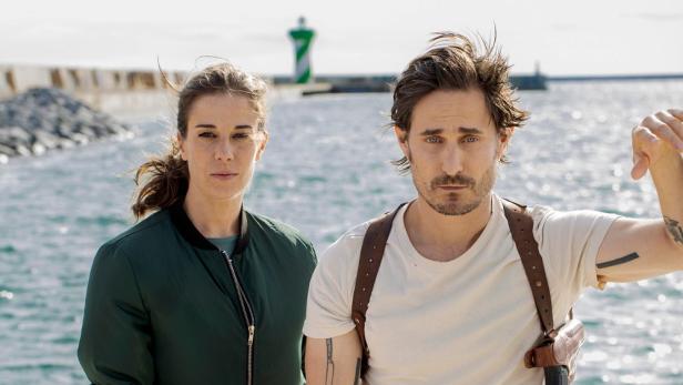 Ein Mann mit Pistole und eine Frau stehen vor einem Leuchtturm im „Barcelona-Krimi“.
