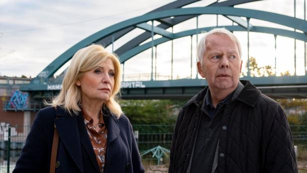 Ein älteres Paar steht vor einer Brücke mit Graffiti im Hintergrund.