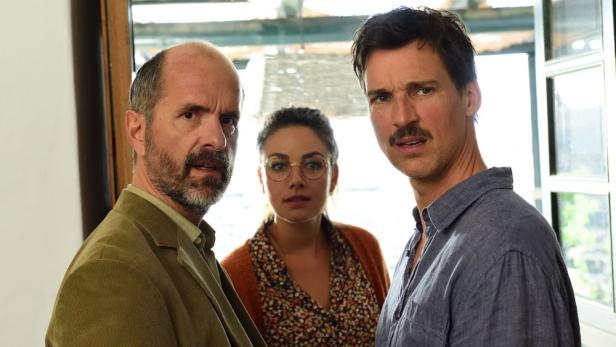 Drei Personen, darunter ein Mann mit Schnurrbart, schauen besorgt in die Kamera.