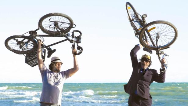 Zwei Männer halten Fahrräder über ihren Köpfen am Strand, während ein Hund zusieht.