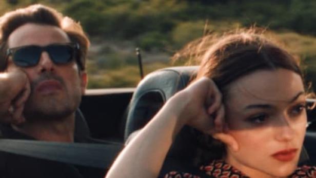 Ein Mann mit Sonnenbrille und eine Frau fahren in einem Cabriolet durch eine grüne Landschaft.