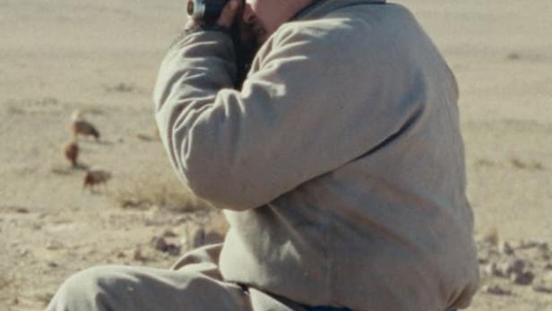 Ein Mann mit Hut sitzt in einer Wüste und beobachtet etwas mit einem Fernglas.