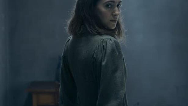 Eine Frau in einem grünen Kleid blickt in einem düsteren Raum über ihre Schulter.