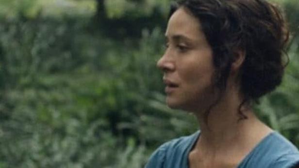 Eine Frau mit dunklen Haaren und blauem Kleid steht vor einer grünen Landschaft.