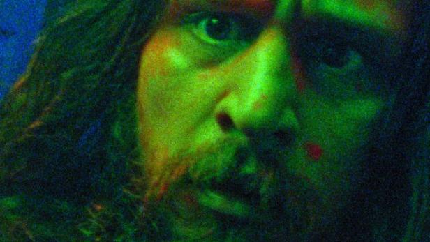 Ein Mann mit Bart und langen Haaren, beleuchtet in grünem Licht, blickt in die Kamera.