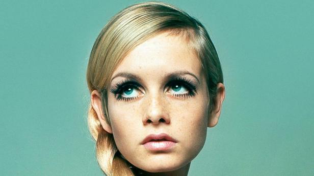 Porträt der jungen Twiggy mit geflochtenem Zopf und auffälligem Make-up vor türkisem Hintergrund.