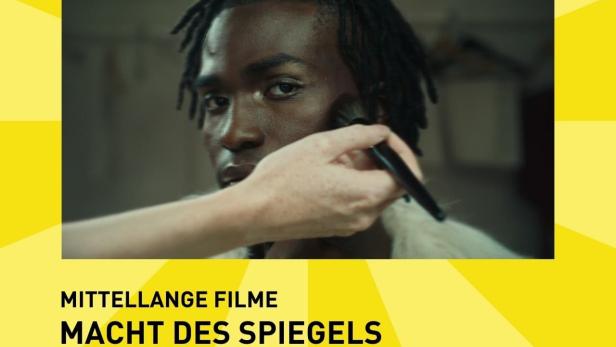 Einem Mann wird Make-up aufgetragen, im Hintergrund das Filmplakat „Macht des Spiegels“.