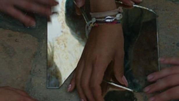 Mehrere Hände berühren zerbrochene Spiegelstücke auf einem Steinboden.