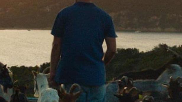 Ein Mann steht inmitten einer Ziegenherde mit Blick auf eine Küstenlandschaft bei Sonnenuntergang.