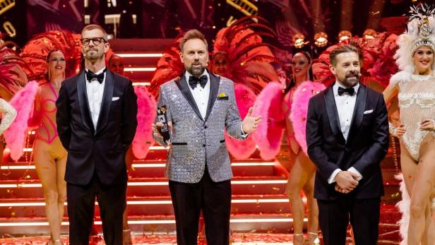 Drei Männer in Anzügen stehen auf einer Bühne, umgeben von Tänzerinnen in Kostümen.