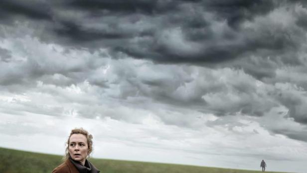 Eine Frau steht auf einem Feld unter einem dramatischen Himmel; im Hintergrund eine weitere Person.