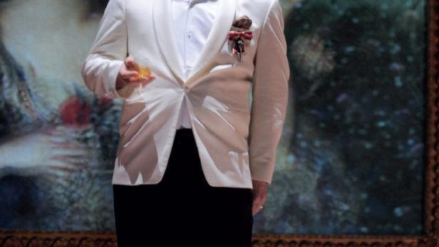 Ein Schauspieler in weißem Anzug steht vor einem Bühnenbild für „Was ihr wollt“.