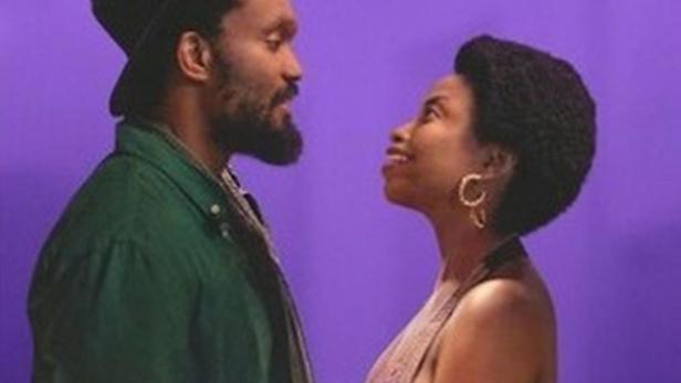 Ein Mann und eine Frau stehen vor einem lila Hintergrund und schauen sich an.