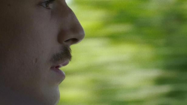 Ein Mann mit Schnurrbart fährt Auto; im Hintergrund verschwimmt die grüne Landschaft.