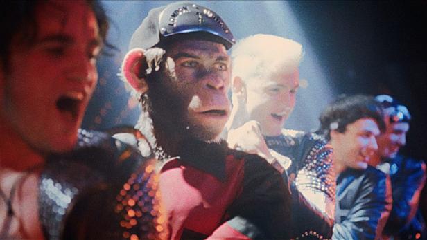 Ein Affe mit einer Londoner Zeitungskappe steht mit anderen in glitzernden Jacken.