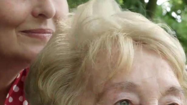 Eine ältere Frau mit blonden Haaren und eine jüngere Frau im gepunkteten Kleid schauen nach oben.