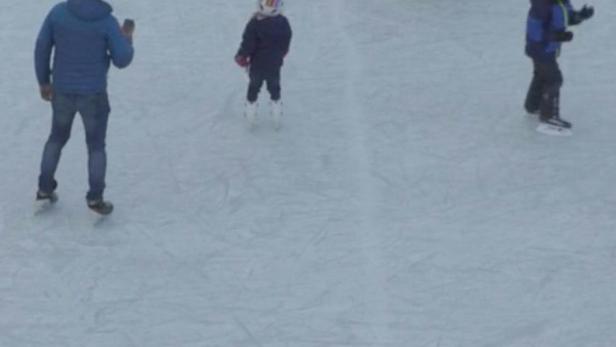 Mehrere Personen, darunter Kinder mit Helmen, laufen auf einer Eisbahn Schlittschuh.