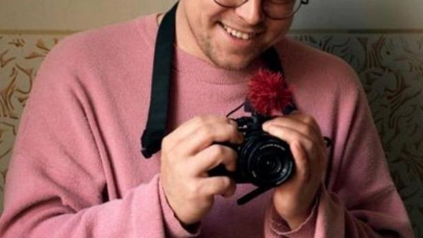 Ein Mann mit Brille und rosa Pullover hält eine Kamera in den Händen. Im Hintergrund hängt ein Porträt von Putin.