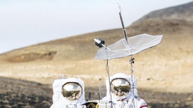 Zwei Astronauten stehen mit einem Rover inmitten einer Schafherde vor einer Hügellandschaft.