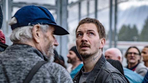 Ein Mann mit Tätowierung am Hals schaut in einer Menschenmenge nach oben.