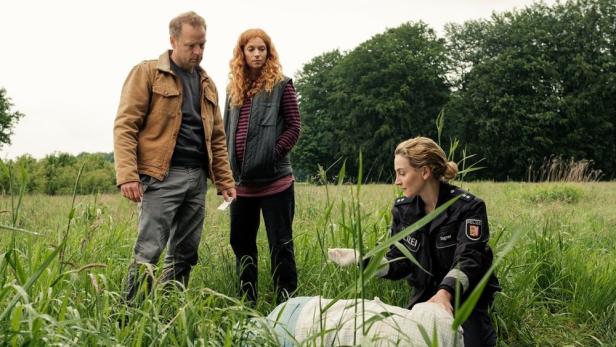 Ein Polizist untersucht eine Leiche in einem Feld, während zwei weitere Personen zusehen.