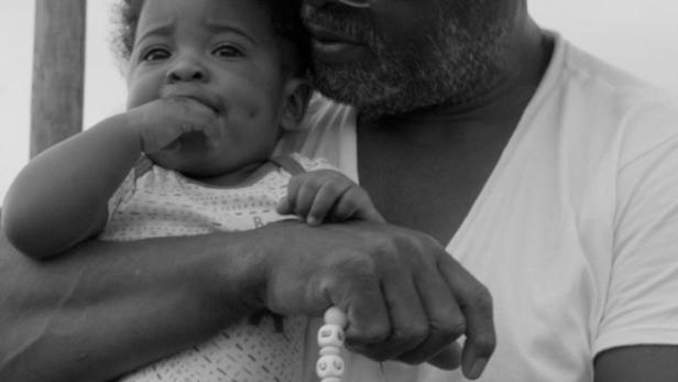 Ein Mann mit Hut und Brille hält ein Baby mit einem Schnuller im Arm.