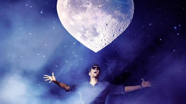 Ein Mann sitzt vor einem herzförmigen Mond und einem Sternenhimmel.