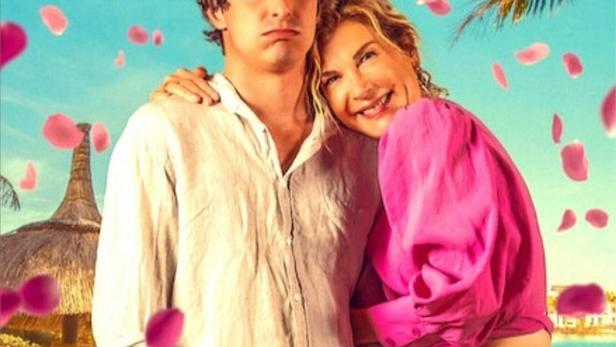 Das Filmplakat für „Honeymoon Crasher“ zeigt ein Paar am Strand, umgeben von Blütenblättern.