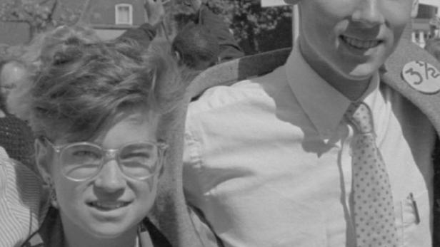 Ein junges Paar lächelt bei einer Demonstration mit einem Schild „Maine“ im Hintergrund.