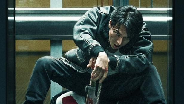 Ein Mann mit einem blutigen Schwert kauert über einer Leiche im Fahrstuhl, im Hintergrund das Logo von „Demon City“.