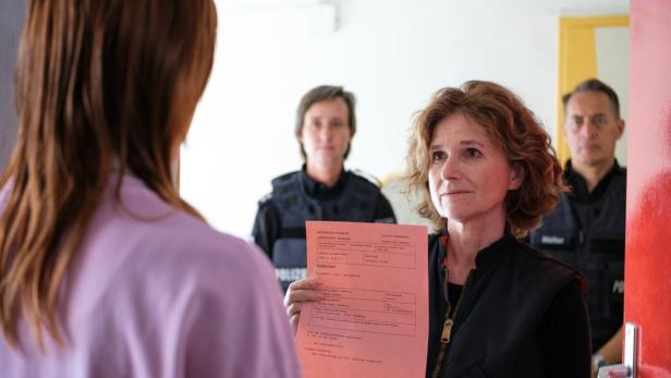 Eine Frau hält einen Haftbefehl vor eine andere Frau, im Hintergrund Polizisten.