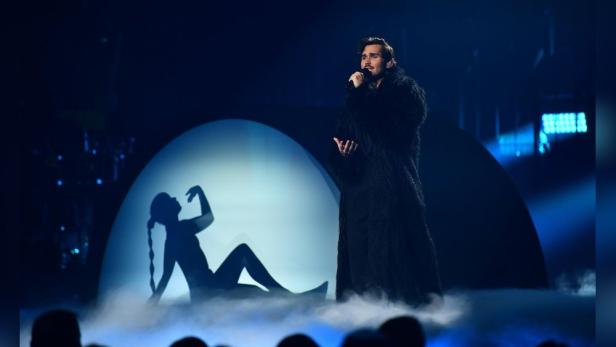 Ein Mann in einem schwarzen Mantel singt auf einer Bühne mit einer Schattenprojektion im Hintergrund.