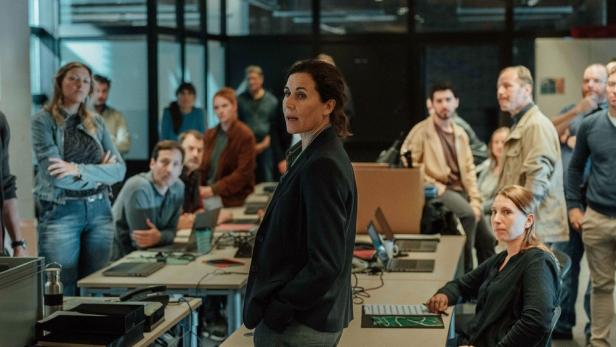 Eine Frau in einem dunklen Blazer spricht vor einer Gruppe von Menschen in einem Büro.