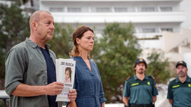 Ein Mann und eine Frau halten ein Vermisstenplakat eines jungen Mädchens in der Hand, im Hintergrund zwei Polizisten.