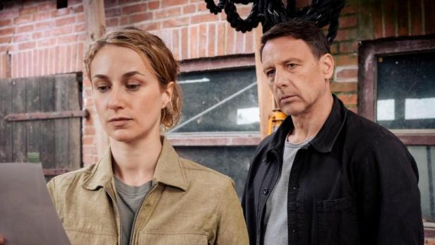 Zwei Personen, vermutlich Ermittler, betrachten ein Dokument in einem Backsteingebäude.