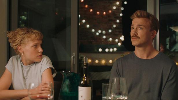 Ein Paar sitzt an einem Tisch mit Wein und Gläsern, im Hintergrund Lichterketten.