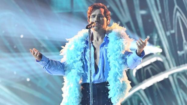 Ein Mann mit blauem Federboa singt auf einer Bühne ins Mikrofon.