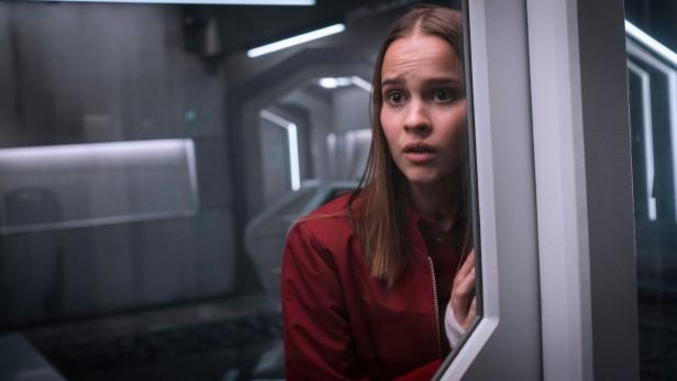 Eine junge Frau mit ängstlichem Blick in einem futuristischen Gang.