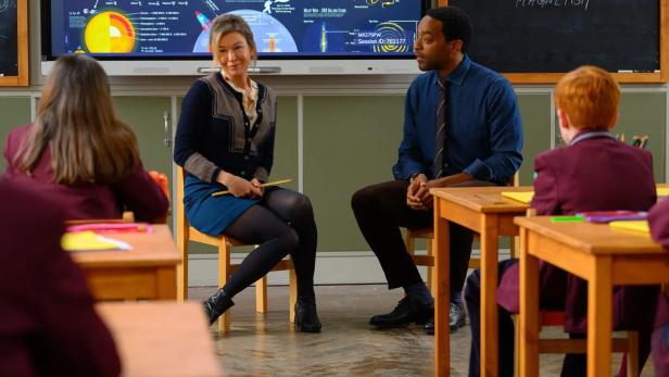 Zwei Lehrer sitzen in einem Klassenzimmer vor einer Tafel mit dem Thema „Infografik“.