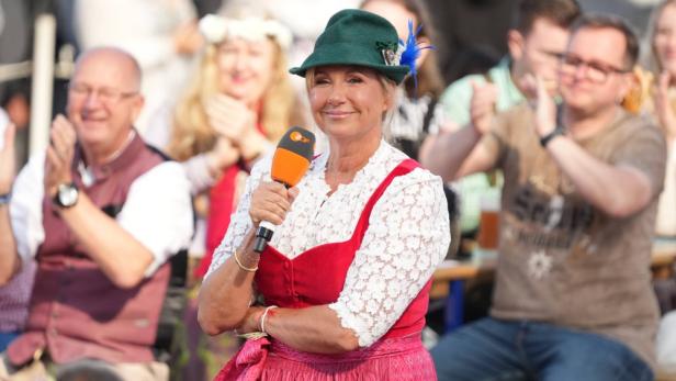 Andrea Kiewel im Dirndl mit Mikrofon beim ZDF-Fernsehgarten, umgeben von applaudierenden Zuschauern.