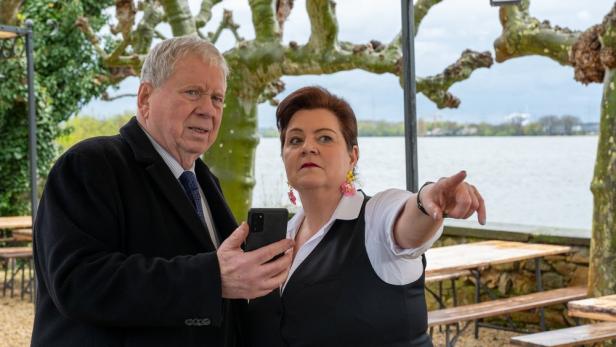 Ein Mann mit Handy und eine Frau zeigen auf etwas am Ufer.