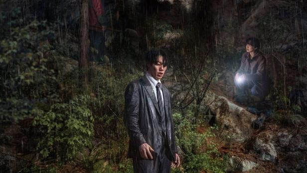 Ein Mann im Anzug steht im Regen, im Hintergrund zwei weitere Personen.