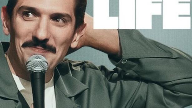Der Comedian Andrew Schulz mit Mikrofon vor dem Cover seines Programms „Life“.