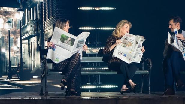 Heidi Klum, Catherine Deneuve und ein Mann sitzen auf einer Bank und lesen Zeitungen.